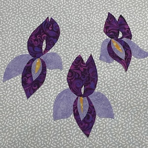 May include: Three stylized iris flower appliqu&eacute;s. The petals are a light purple, with a darker purple, patterned center. The center of each flower is yellow. The background is a light gray fabric with a white leaf pattern.
