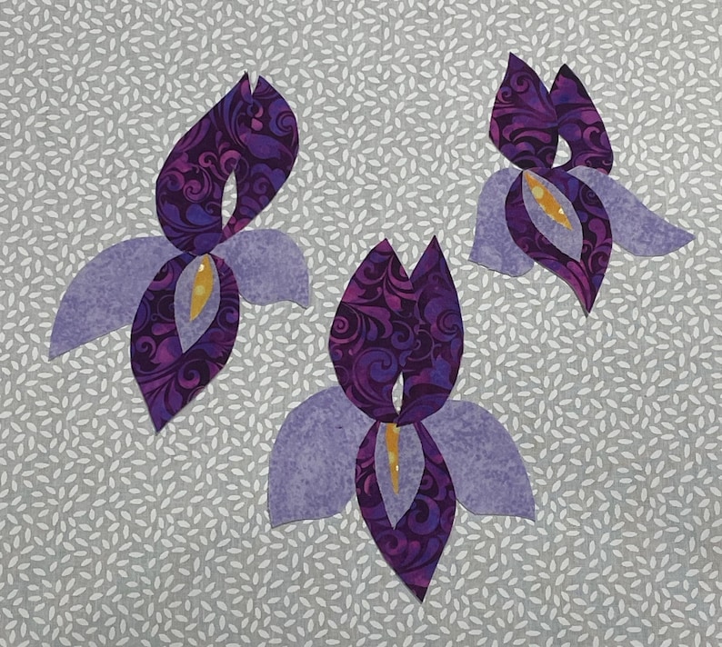 May include: Three stylized iris flower appliqu&eacute;s. The petals are a light purple, with a darker purple, patterned center. The center of each flower is yellow. The background is a light gray fabric with a white leaf pattern.