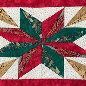May include: A red, green and gold patchwork quilt block featuring a star design. The star is constructed from different fabric scraps sewn together in a geometric pattern.