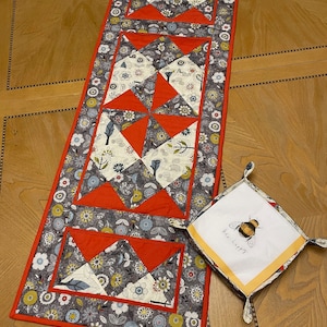 以下が含まれることがあります： 手作りのキルトテーブルランナーと、お揃いの正方形のトレイ。ランナーは、赤とグレーのアクセントのフローラルパターンが特徴です。トレイには、蜂のグラフィックと「bee happy」の文字が描かれています。