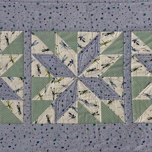 May include: A rectangular quilt with three star-shaped blocks. Each star is made from light blue, green, and cream fabrics, patterned with dragonflies. The quilt has a light blue speckled border.