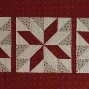 May include: Three square quilt blocks with a star pattern. Each block features a central eight-pointed star design in red and white, with patterned triangles in the corners. The background is a solid, dark red fabric.