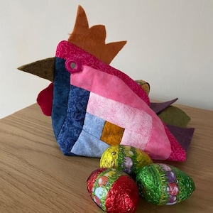 Peut inclure: Un poulet en tissu patchwork coloré avec un bec en feutre brun et une crête en feutre orange. Le poulet tient trois œufs de Pâques en chocolat enveloppés de papier d'aluminium dans ses ailes.