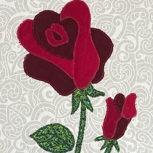 May include: A fabric applique featuring a large, deep red rose and a smaller rosebud. The petals are crafted from layered red and dark maroon felt with visible blanket stitching. The stems and single leaf are made of green textured fabric.