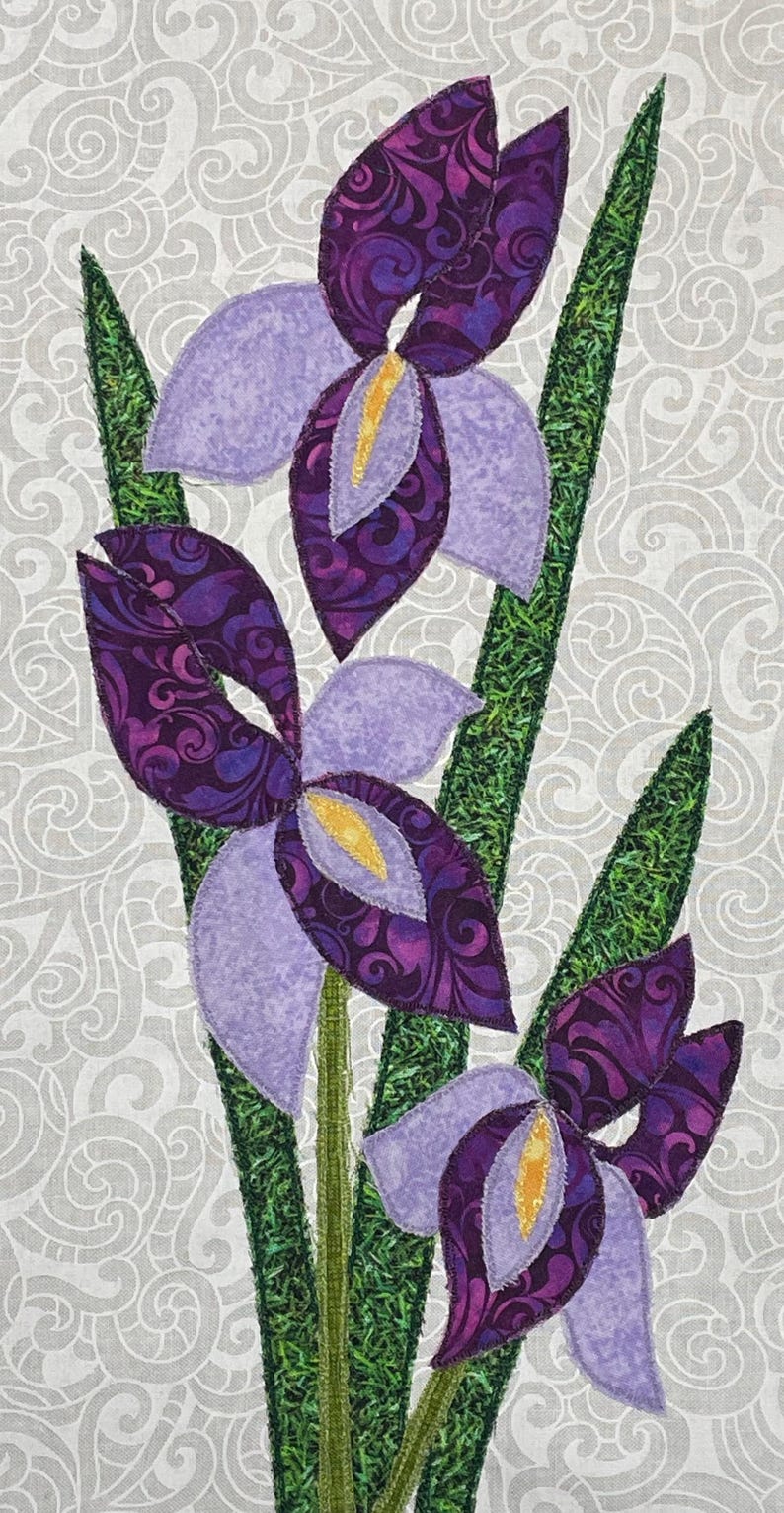 May include: A fabric art piece featuring three stylized iris flowers. The flowers have purple petals with patterned centers and yellow accents. Green stems and leaves are visible against a light gray background with a swirling pattern.