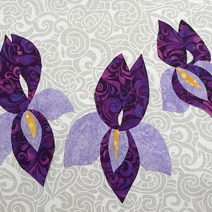 May include: Three fabric iris flower cutouts in shades of purple, lavender, and yellow. The petals are a solid lavender, while the inner petals feature a swirling pattern of deep purple. The background is a light gray fabric with a swirling design.