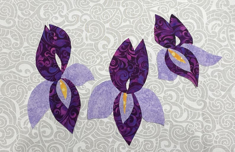 May include: Three fabric iris flower cutouts in shades of purple, lavender, and yellow. The petals are a solid lavender, while the inner petals feature a swirling pattern of deep purple. The background is a light gray fabric with a swirling design.