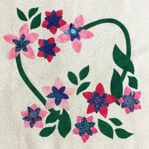 May include: A green vine with pink and red flowers with blue centers. The flowers are made of fabric and are appliqued onto a white background.