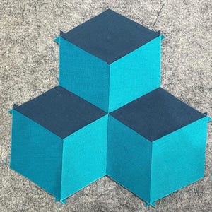 Op de afbeelding: Geometrisch stoffkunstwerk met drie verbonden kubusvormen. De bovenkanten zijn donker marineblauw en de zijkanten helder turquoise. Het werk ligt op een grijze ondergrond.