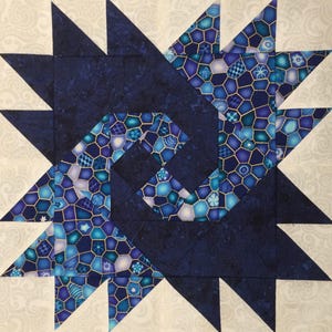 May include: A blue and white quilt block with a geometric pattern. The block is made up of several smaller squares and triangles, and the pattern is created by the different colors and shapes of the pieces. The block is surrounded by a white background.