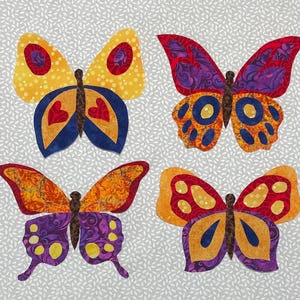 May include: Four colorful butterfly fabric appliqués. Each butterfly has unique wing patterns in shades of yellow, orange, red, purple, and blue. The butterflies are set against a light gray background with a subtle leaf pattern.