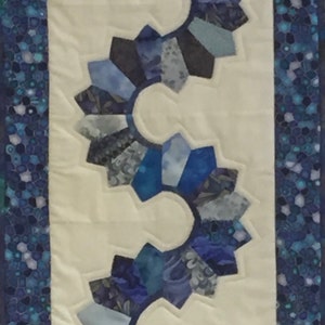May include: A white fabric table runner with a blue and grey patchwork design. The design features a series of connected circles with a scalloped edge.
