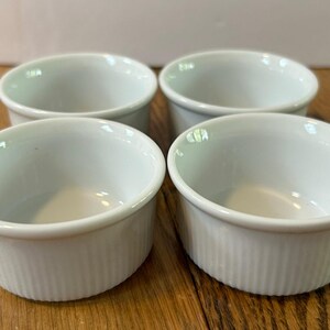 Puede incluir: Cuatro ramequines blancos y redondos con lados acanalados verticales. Los ramequines están dispuestos en un cuadrado sobre una superficie de madera. El interior de los ramequines es liso y blanco.