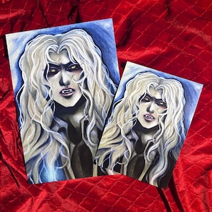 May include: Two art prints depicting a character with long, flowing white hair, yellow eyes, and fangs. The character is set against a blue backdrop. The prints are displayed on a red velvet surface.