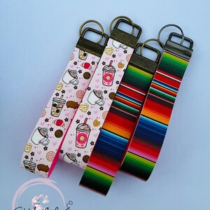 May include: Four keychains with bronze-colored hardware. Two keychains feature a pink background with coffee and pastry illustrations. The other two have colorful striped patterns, one with a serape design. The keychains are on a light blue background.