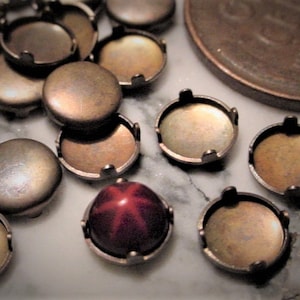 May include: A collection of antique bronze-colored bezel settings and cabochon settings. The settings are round with a flat base and a raised edge to hold a stone or other insert. One setting holds a red star-shaped stone.