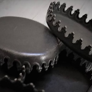 May include: A collection of matte black metal bottle caps featuring a smooth, flat circular top and a serrated, scalloped edge design. These crown-style caps are arranged in a cluster, showcasing their dark, uniform finish and distinct, jagged rim structure.