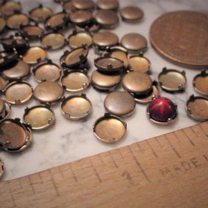 May include: A collection of small, antique brass-colored bezel settings and domed metal components. A red, star-shaped cabochon is set in one bezel. A wooden ruler with "MADE IN U.S." is in the frame.
