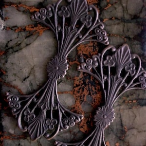 May include: Two ornate, silver-toned metal decorative elements. Each features a central floral design with radiating branches of stylized leaves and small, round accents. The pieces are set against a mottled green and brown background.