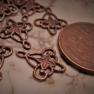 May include: Several copper-colored metal filigree connectors featuring a four-pointed star design with looped edges. Each connector has two small circular loops for attachment. A United States one-cent coin is placed next to the metal pieces.