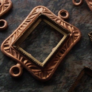 May include: A copper-toned metal frame with an ornate design and a rectangular opening for a cameo or other embellishment. The frame has two loops on the side for attaching to a necklace or bracelet.
