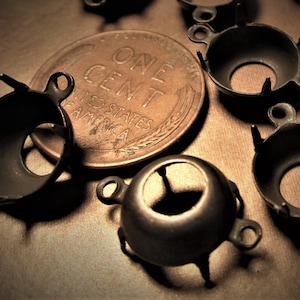 May include: A close-up of a collection of antique-looking metal settings for jewelry making. The settings are round with a small hole in the center and have four prongs on the bottom. The settings are arranged on a brown surface next to a US penny.