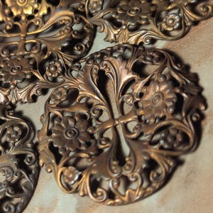 May include: Close-up of several ornate, bronze-colored metal filigree pieces. The designs feature floral and leaf patterns with intricate details. The pieces are arranged on a neutral-toned surface, showcasing their antique aesthetic.