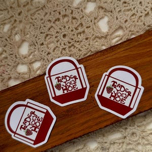 May include: Three stickers with a red and white design. Each sticker has the text "Trader Joe's Hoe" and a strawberry graphic. The stickers are on a wooden surface with a cream-colored crocheted background.