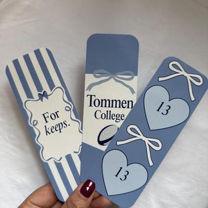 May include: Three blue and white bookmarks with different designs. One has vertical stripes and the text "For keeps." Another says "Tommen College" with a bow graphic. The third has two hearts with the number "13" and bow graphics.