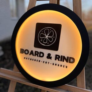 Pode incluir: Uma placa redonda com uma borda preta e um interior quente e brilhante. A placa apresenta as palavras "BOARD & RIND" em negrito, com "GATHERED - CUT - SHARED" abaixo. Um gráfico quadrado com um design cítrico está no topo.