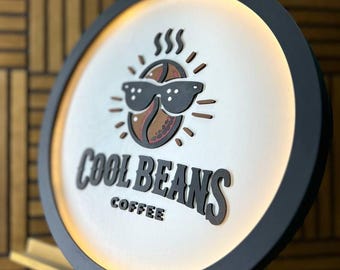 May include: A round sign with a black frame and a white interior. The sign features a coffee bean wearing sunglasses, with the words "COOL BEANS COFFEE" in a bold font. The sign is illuminated with a warm light.