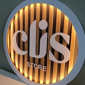 Puede incluir: Un letrero redondo con marco blanco y fondo de listones de madera. El letrero tiene la palabra "clis" en letras blancas y la palabra "store" en letras blancas más pequeñas debajo. El letrero está iluminado desde atrás, creando un brillo cálido.