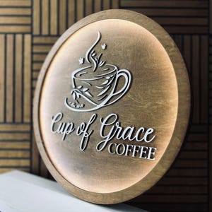 May include: A round wooden sign with the words "Cup of Grace COFFEE" in white script. The sign features a white coffee cup design with steam and leaves. The sign has a warm, glowing light around the edge.