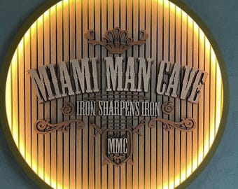 Op de afbeelding: Een rond houten bord met een gouden rand en een verlichte halo rond de rand. Het bord zegt "Miami Man Cave" met de woorden "Iron Sharpens Iron" eronder. Het bord heeft ook de letters "MMC" in een schildvorm.