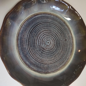 May include: A ceramic plate with a dark brown and blue glaze. The plate has a wavy rim and a spiral pattern in the center. The plate's color transitions from dark brown to blue and white.