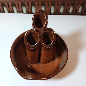 May include: A brown ceramic vase with three cylindrical openings and a shallow dish base. The vase has a speckled glaze and a unique, organic shape. The color is a warm, earthy brown, suitable for holding flowers or other decorative items.