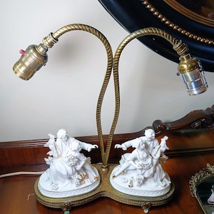 May include: Antique brass table lamp with two curved arms. The lamp features two white porcelain figurines of a couple playing musical instruments. The base is a decorative brass with ornate detailing. The lamp has two light sockets.