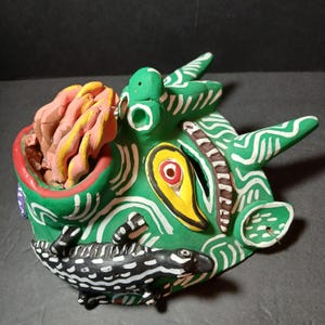 May include: A handcrafted, colorful ceramic mask with a green base, white accents, and a black and white reptile figure. The mask features a red, yellow, and pink design, with a detailed, textured appearance. The mask is a unique piece of art.