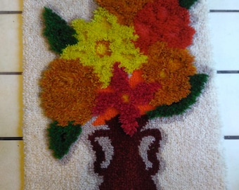 May include: A rectangular, textured wall hanging featuring a floral design. The flowers are in shades of yellow, orange, and red, with green leaves, emerging from a dark maroon vase. The background is off-white, with a brown border.