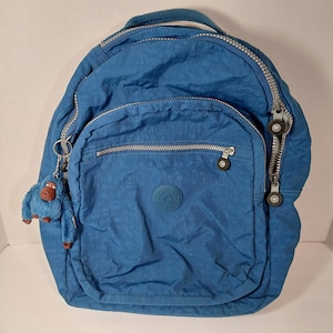 May include: A blue backpack with multiple zippered compartments and a top handle. The backpack features a small monkey keychain and a circular logo on the front. The zippers are silver-toned, and the material has a slightly wrinkled texture.