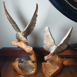 May include: Two wooden eagle sculptures with outstretched wings. The sculptures are carved from light brown wood and are perched on stylized wooden bases. The eagles' wings and feathers are intricately detailed, showcasing the craftsmanship of the carvings.