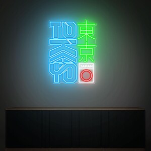 May include: Neon sign with the word "TOKYO" in blue and green, with Japanese characters. The sign includes a red and white circle with the word "JAPAN" below it. The sign is mounted on a dark wall above a dark cabinet.