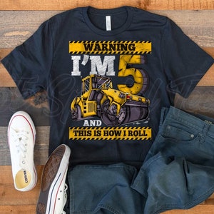May include: Dark navy blue t-shirt with a yellow and black construction-themed graphic that reads "WARNING I'M 5 AND THIS IS HOW I ROLL." The shirt is paired with blue jeans and white canvas sneakers.