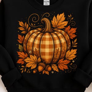 May include: Black sweatshirt with a plaid pumpkin design in orange and yellow, surrounded by autumn leaves and acorns. The graphic is centered on the front, evoking a fall harvest theme. The Gildan brand label is visible.
