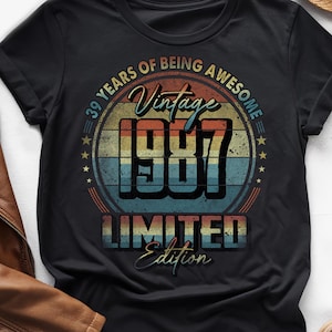 Puede incluir: Camiseta negra con un diseño vintage que celebra un 39 cumpleaños. El diseño presenta el texto "39 Years of Being Awesome Vintage 1987 Limited Edition" en una combinación de colores retro naranja, azul y amarillo.