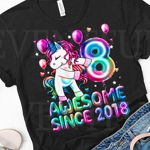 Peut inclure: T-shirt noir avec un motif de licorne colorée, avec une corne bleue et une crinière rose, faisant un dab à côté d'un grand ballon en forme de chiffre 8. Le texte dit "AWESOME SINCE 2018". Ballons et étoiles complètent le design.