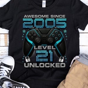 May include: Black t-shirt with a gaming theme. The shirt features the text "AWESOME SINCE 2003" above a teal and black game controller graphic. Below the controller, the text reads "LEVEL 21 UNLOCKED".