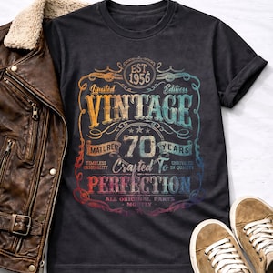 Könnte beinhalten: Dunkelgraues T-Shirt mit farbenfrohem Vintage-Design. Das Design enthält den Text "EST. 1956", "VINTAGE", "70 YEARS" und "Crafted to Perfection". Eine braune Lederjacke und beige Sneaker sind ebenfalls im Bild.