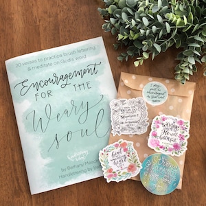 May include: A book titled "Encouragement for the Weary Soul" by Bethany Meacha with hand lettering by Beth. The book cover is a light blue watercolor background with white lettering. The book is packaged with a brown paper envelope containing four stickers with floral designs and inspirational quotes. The quotes include "I know that you can do all things...", "Bind up the brokenhearted...", "The Savior loves me so...", and "Be strong and courageous..."