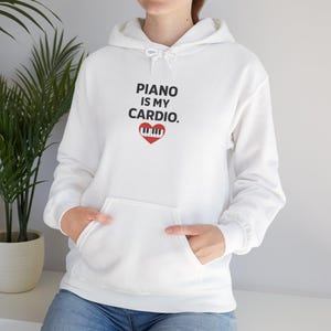 Peut inclure: Un sweat à capuche blanc avec une poche avant et un cordon de serrage. Le texte "PIANO IS MY CARDIO" est imprimé en noir au-dessus d'un cœur rouge avec un motif de clavier de piano. Le sweat à capuche est en matière douce.